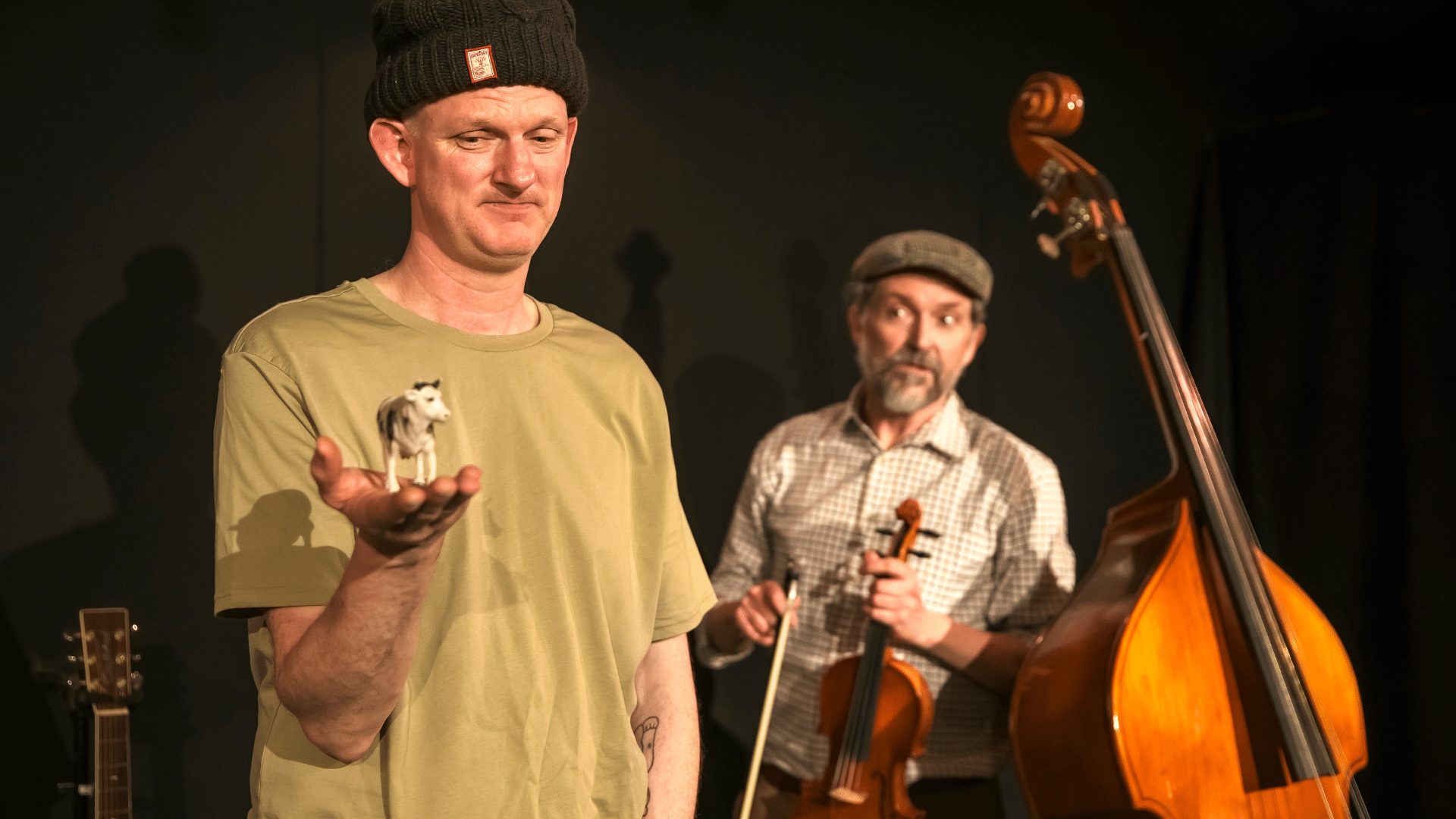 One Man and His Cow production photo - On the left in the foreground: Stu McLoughlin holding a miniature cow figurine in the palm of his right hand, as he looks down on it. He wears a black beanie hat with his ears sticking out, and a cream coloured t-shirt. In the background, to the right: Ian Harris looks on at Stu while holding a violin in his left hand and the bow in his right hand. To his left, a double-bass is stood up. Ian wears a farmer-style flat cap and a white square-patterned shirt. Background: black stage curtain.