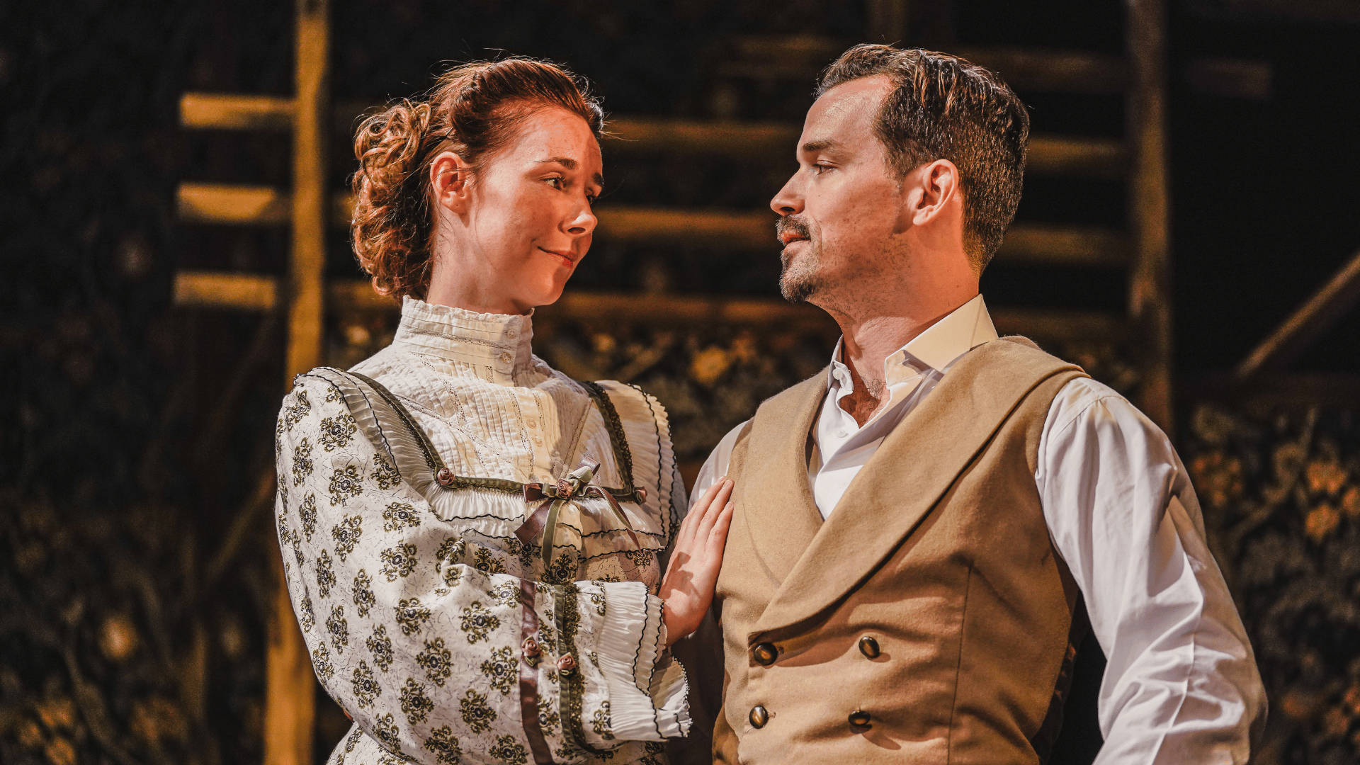 A woman in a Victorian dress with her hand on the arm of a man a tan coloured waistcoat. They stare into each other's eyes.