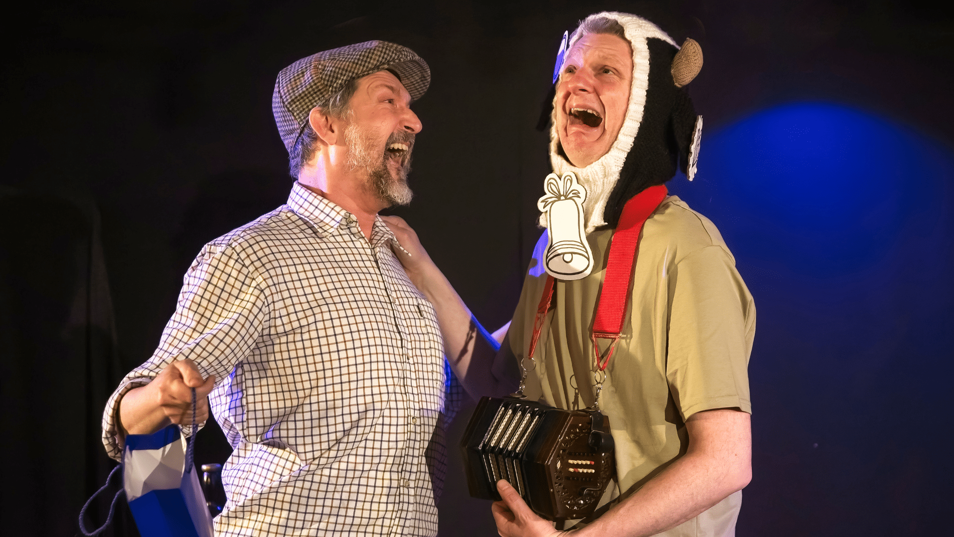 One Man and His Cow production photo - Ian Harris (left) and Stu McLoughlin (right) stood together with an arm each on one another's shoulder. They both have their mouths open as if yelling and Ian faces Stu, while Stu looks upwards, facing his right. Ian wears a farmer-style flat cap and a white square-patterned shirt and holds a blue gift bag in his right hand. Stu wears a knitted balaclava hat with an opening big enough for his full face and patterned black and white like a cow with small cow horns sticking out from the top. He also wears a cream-coloured t-shirt, a cardboard cow bell and an accordion on a strap around the back of his neck. Background: black stage curtain with blue spot light shining on it on the right.