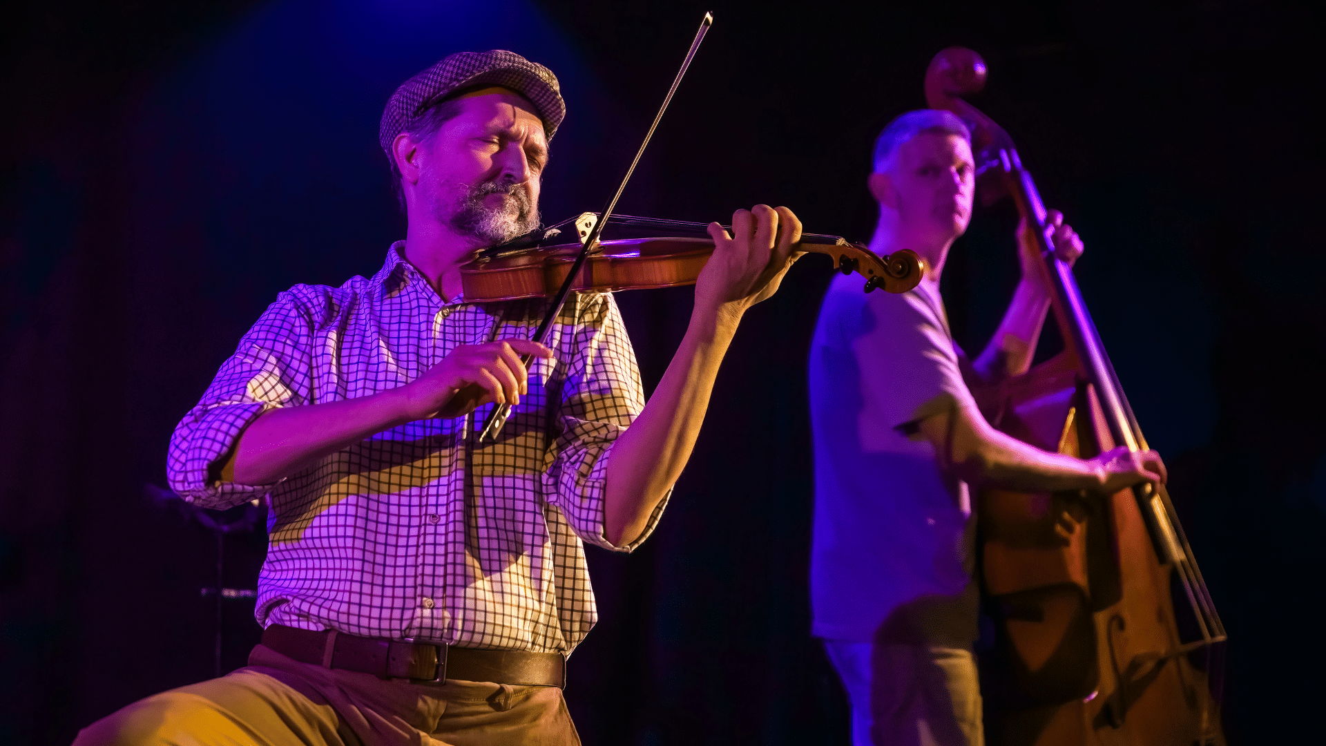 One Man and His Cow production photo - On the left, in the foreground: Ian Harris playing the violin. His eyes are closed in concentration and he wears a farmer-style flat cap and a white square-patterned shirt. On the right, in the background: Stu McLoughlin plays the double-bass. Background black with blue light.