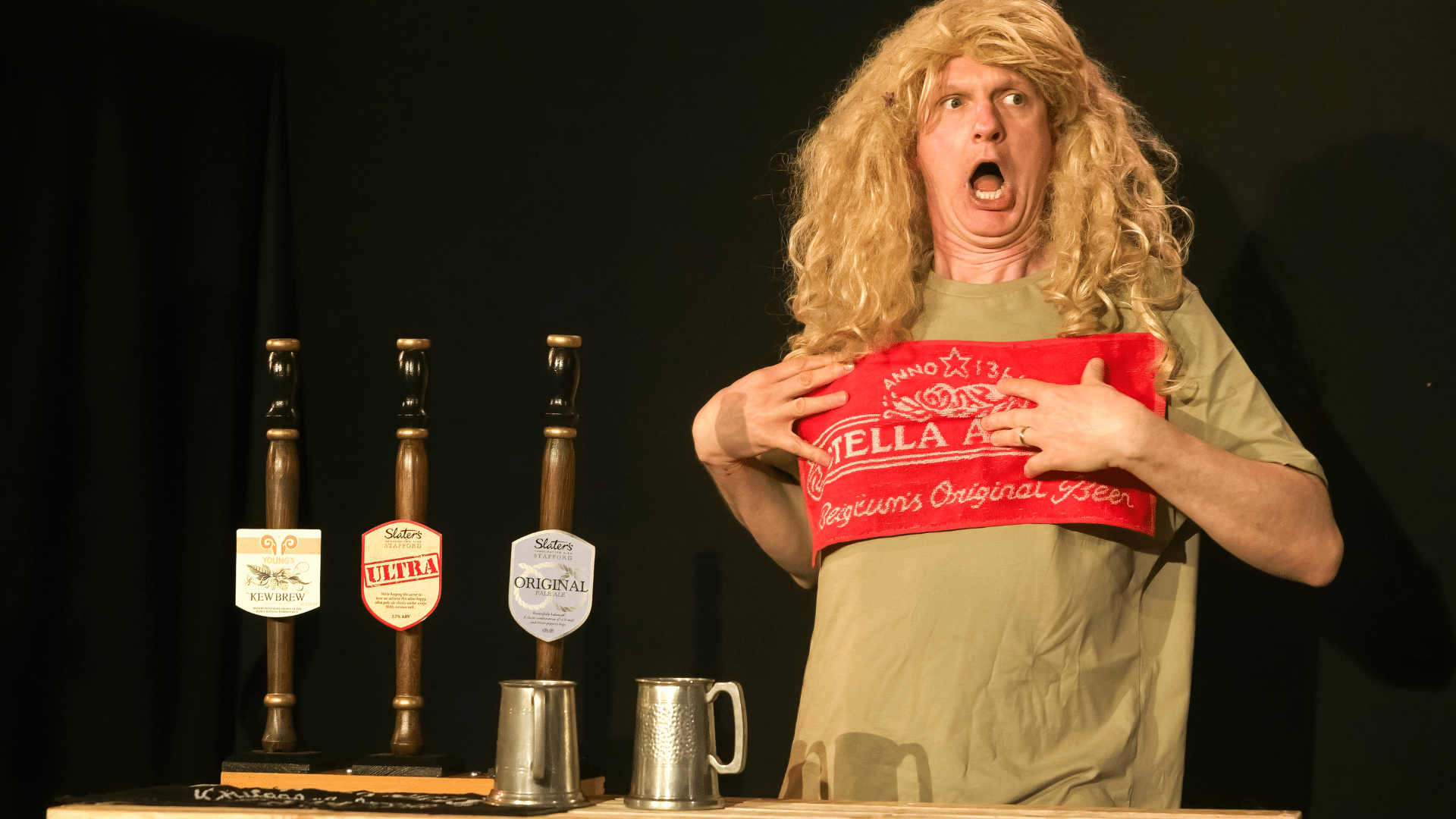 One Man and His Cow production photo - Stu McLoughlin (right) holding a bar cloth across his chest with his mouth open in an expression of shock. He wears a blonde long-haired wig and a cream-coloured t-shirt. On his left are three beer pumps and two pewter drinking tankards on a bar top. Background: black stage curtain