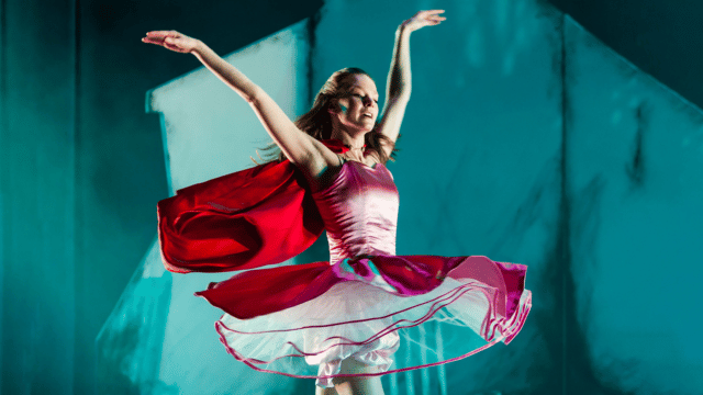 A young dancer in a pink flowy dress with a red cape is doing a pirouette on pointe, arms elegantly lifted in the air
