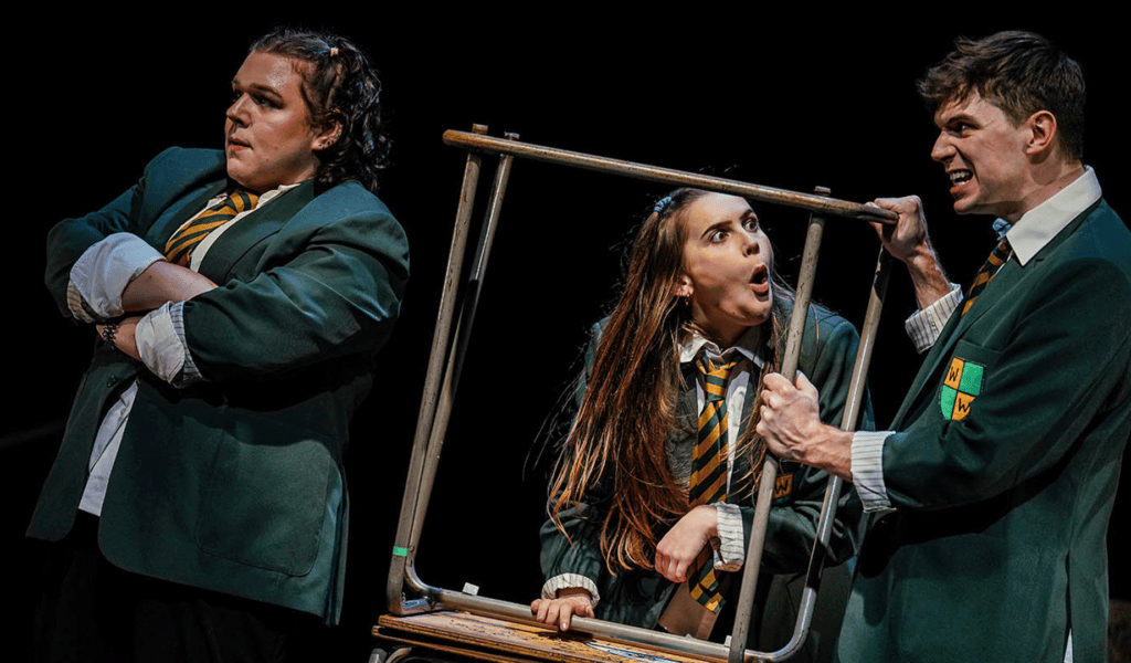 Teechers Leavers ’22 production image. Background: A theatre stage. Black curtain backdrop. Foreground: (left) A young woman, wearing a green school blazer, white shirt and green and yellow tie, crosses their arms and looks off to the left-hand side of the image. To their right, a young woman (centre) and a young man (right), wearing green school blazers, white shirts and green and yellow ties, argue while holding two school tables positioned on top of each other.