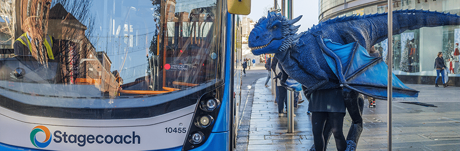Background: Exeter high street. A road and pavement with grey slab tiling stretches to the horizon. Foreground: (Left) a Stagecoach-branded blue and white double decker bus. (Right) A large blue dragon puppet, with horns, a ridged back and light blue winds. The dragon is standing as if about to board the bus.