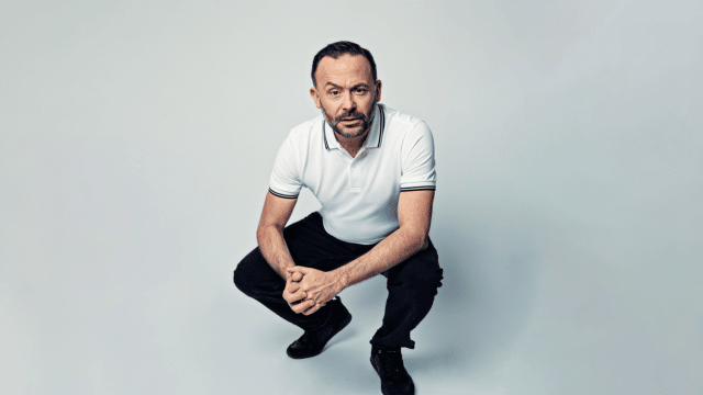 A picture of Geoff Norcott wearing a white polo shirt and black trousers, crouching down, clasping his hands together and facing forwards with an eyebrow arched.