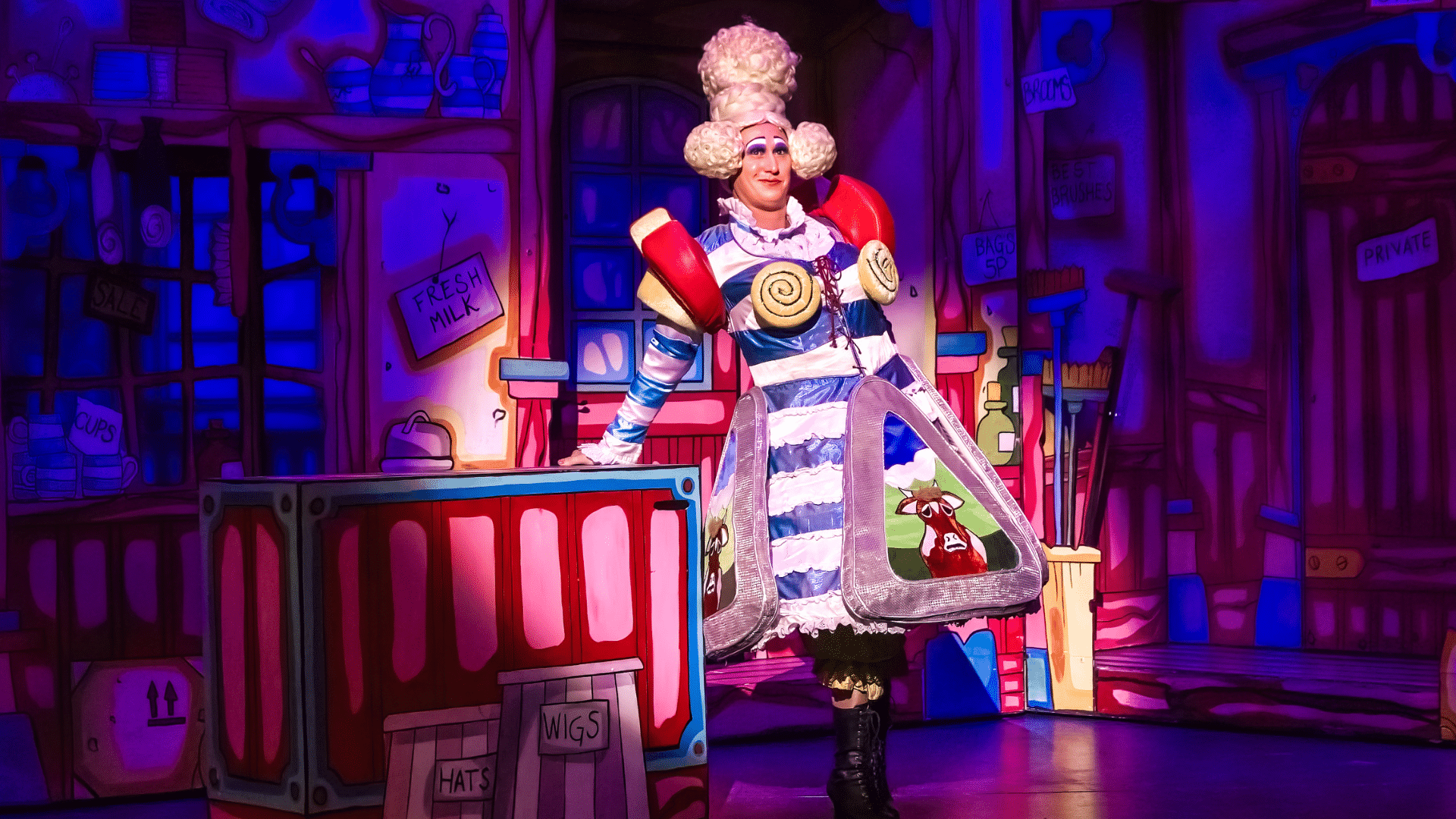 Dick Whittington Production Photo: Sarah Suet (Matt Freeman) leans against a counter in a colourful shop-interior, wearing a globular blonde wig and a dress adorned with cheese triangles.