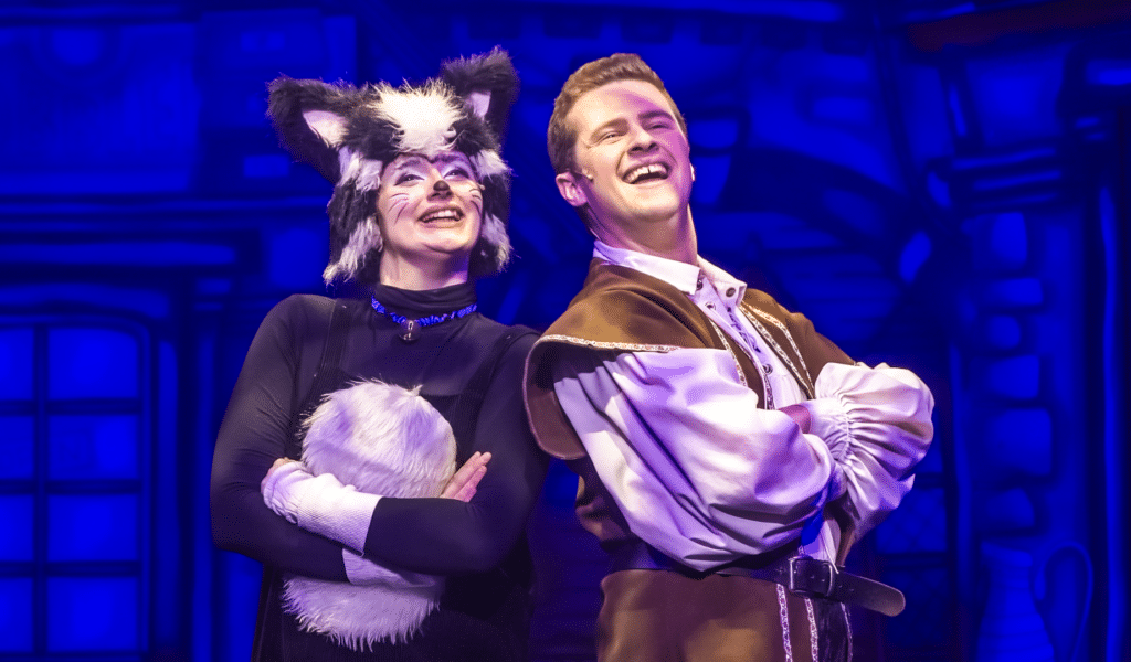 Dick Whittington Production Photo: Dick Whittington (Elliot Coombe) wears a loose white shirt and brown waistcoat; he stands back-to-back with Sox (Poppy Joy), a young woman dressed as a black-and-white cat; they are both crossing their arms.