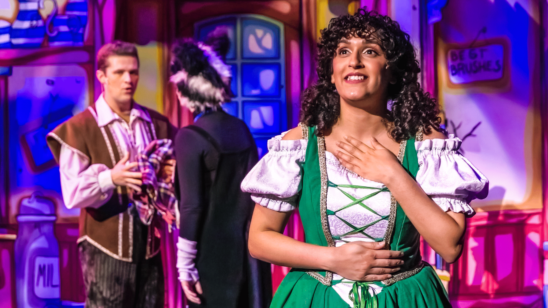 Dick Whittington Production Photo: Sophia Lewis stands in the foreground, wearing a green pinafore dress over a white off-the-shoulder blouse; her hands are resting against her collar bone and abdomen; in the background, Dick (Elliot Coombe) and Sox (Poppy Joy) are in conversation in a colourful shop-interior.