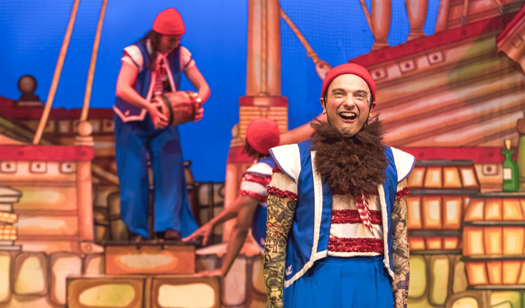 Dick Whittington production photo: Jack (Al Dunn) stands in the foreground of a ship set dressed as a sailor, with tattoo sleeves and bushy fake beard; he is pulling a buck-toothed expression and squinting.