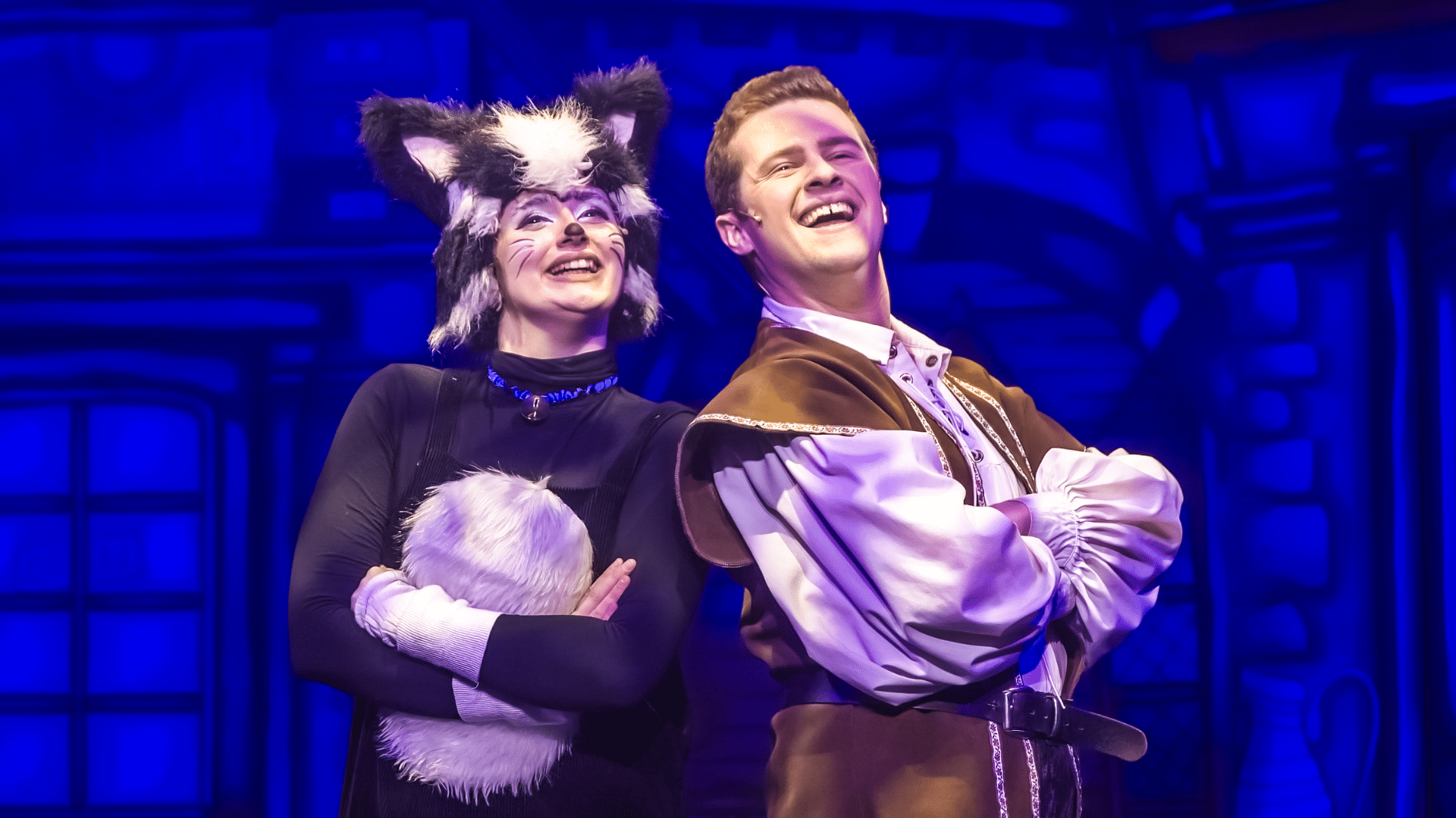 Dick Whittington Production Photo: Dick Whittington (Elliot Coombe) wears a loose white shirt and brown waistcoat; he stands back-to-back with Sox (Poppy Joy), a young woman dressed as a black-and-white cat; they are both crossing their arms.