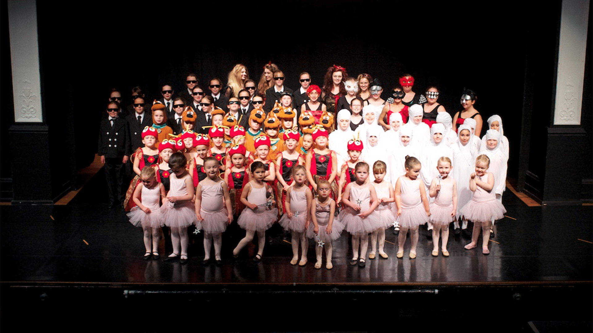 Topsham School of Dance promotional image. Background: The Barnfield Theatre stage. Black backdrop sided by two white pillars. A wooden stage. Foreground: A mixed group of young dancers wearing a variety of fancy dress costumes.