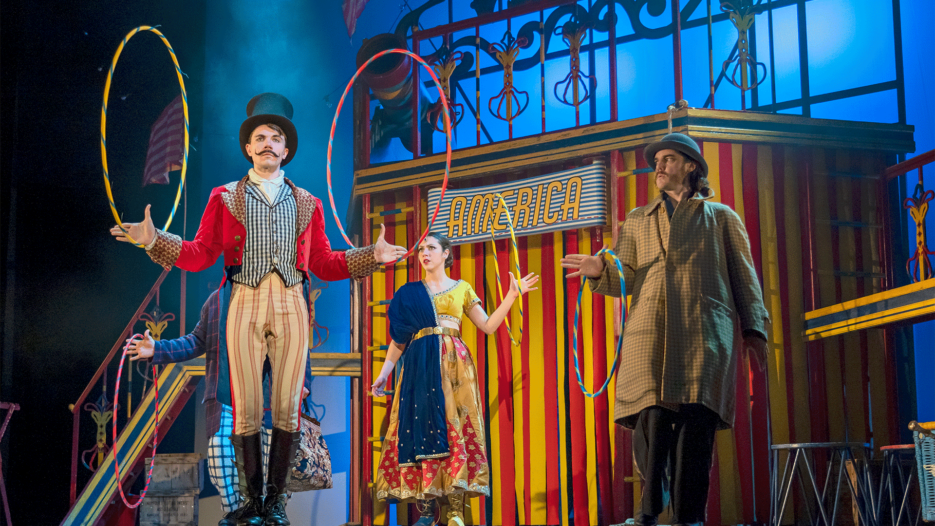 Around the World in 80 Days production image. Background: Blue lights wash through haze. A large red and yellow coloured stripey circus tent structure, with a canon on top and wooden blue, red and yellow coloured ladders leaning on it from all sides. On either side of the circus tent, steps leading up to the top of the structure. Foreground: (left to right) A man, wearing a top hat, red circus ringmaster jacket, checked black and white waistcoat, brown stripey trousers and black boots, twirls two hula-hoops; a woman, wearing a blue, red and yellow dress, twirls a hula-hoop in her left hand; a man, wearing a bowler hat, large beige coat and dark brown trousers, struggles to twirl a hula-hoop with their right hand.
