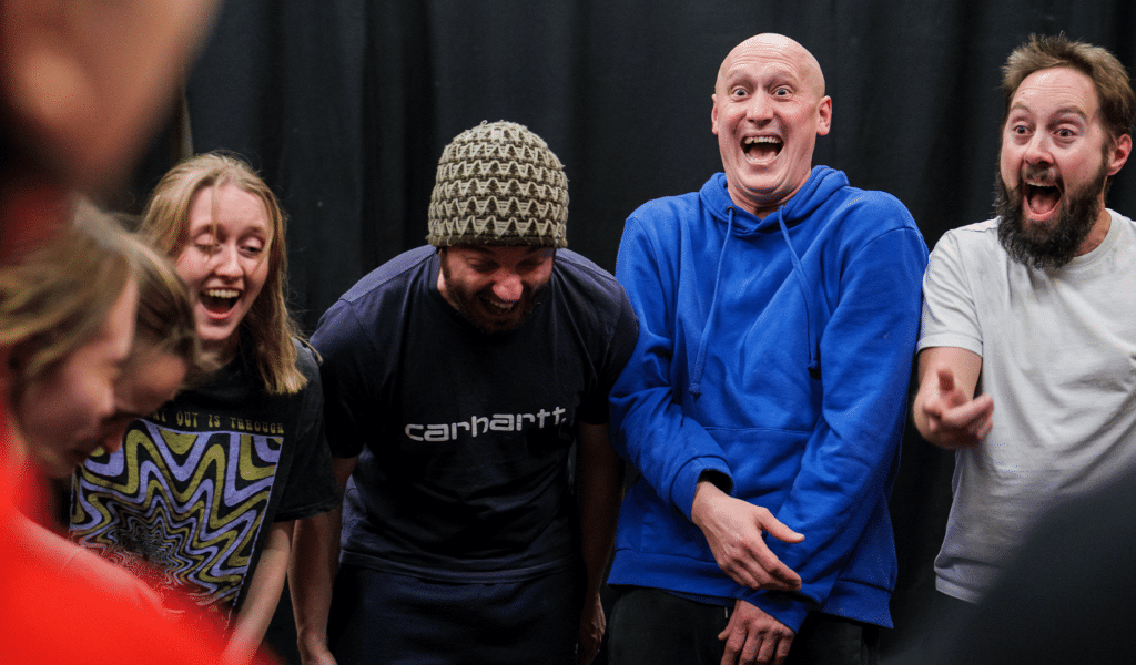 Promotional photo captured during Northcott Futures Physical Theatre Masterclass. Background: Barnfield Clifford Room. Black curtain. Foreground: Al Dunn, Matt Freeman and Nick Bunt (centre to right) laugh as part of a circle made with other performers (left).