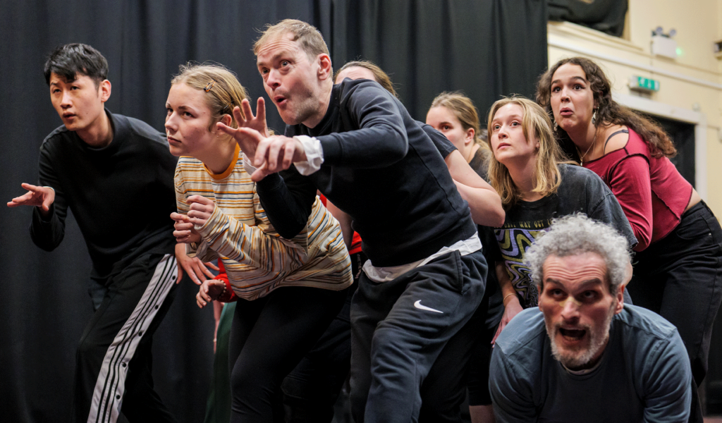 Promotional photo captured during Northcott Futures Physical Theatre Masterclass. Background: Barnfield Clifford Room. A long black curtain draped three-quarters of the way across the photo, (left to right), covering a cream coloured wall. To the right of the image, a black door. Foreground: A group of performers creep past the camera, looking afraid of what's ahead of them.
