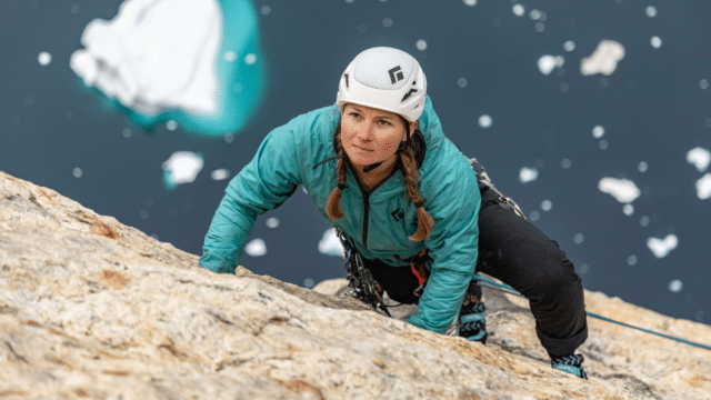 Hazel Findlay (a white woman with long light brown hair) climbing a sheer looking rock face. She wears a white helmet and turquoise waterproof jacket. The camera angle looks down on her as she climbs. Behind/beneath her, we can see dark blue water with large chunks of ice floating on the top.
