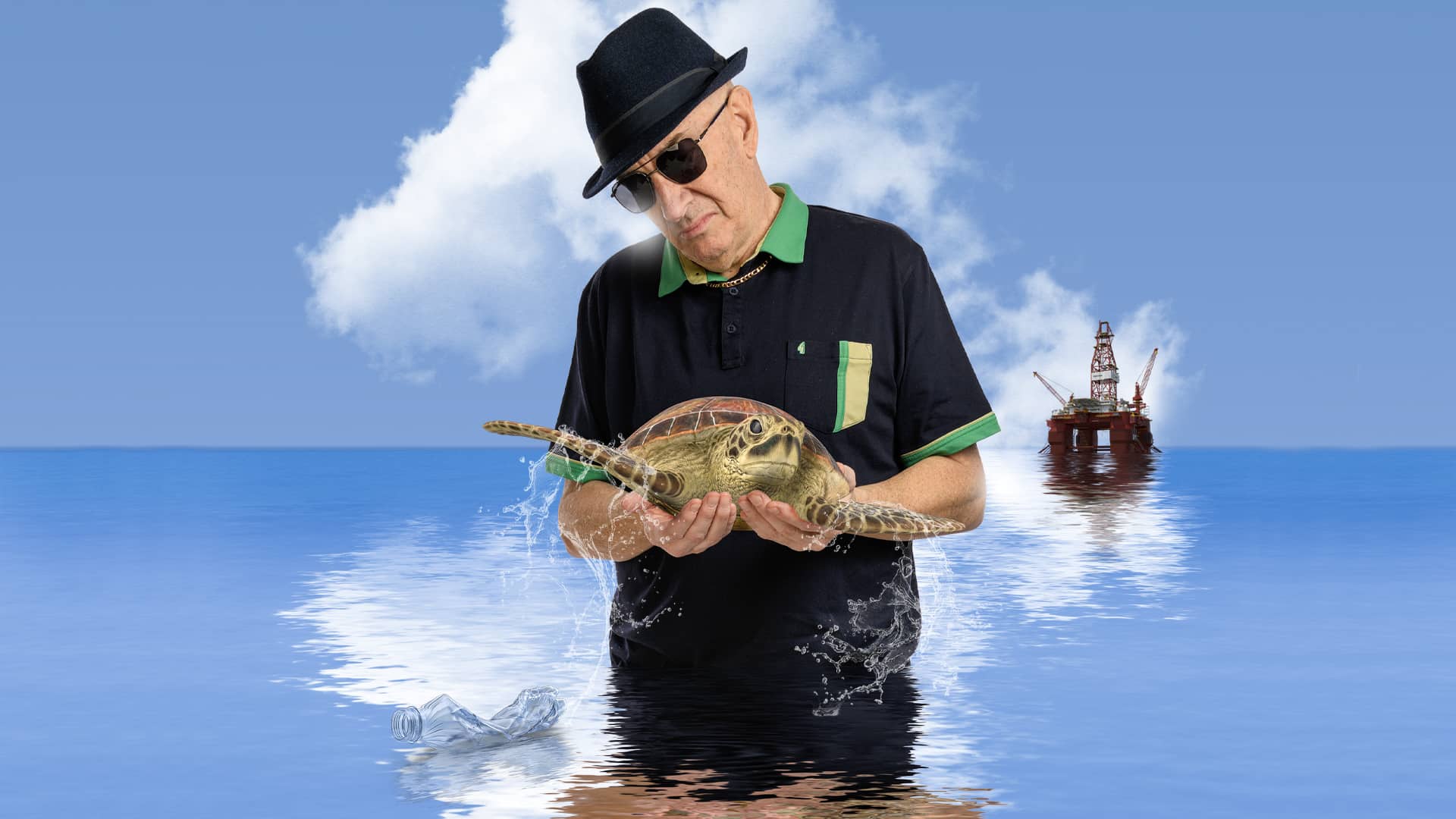 Simon Day as Dave Angel, Eco Warrior - A man in his sixties wears a black trilby hat, dark sunglasses and a black polo shirt with a green collar and sleeve edges. He is standing in the sea and holding a sea turtle. On his right there is a squashed empty plastic bottle floating on the water and behind him to his left is an oil rig in the distance. The sky in the background is blue with some white clouds and the sea water reflects the blue sky.