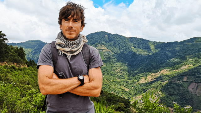 Simon Reeve – To the Ends of the Earth promotional photo. Background. A mountain valley covered with green foliage. Cloudy sky. Foreground: Simon Reeve, wearing a white scarf, grey t-shirt and olive-green trousers, stands with his arms crossed.