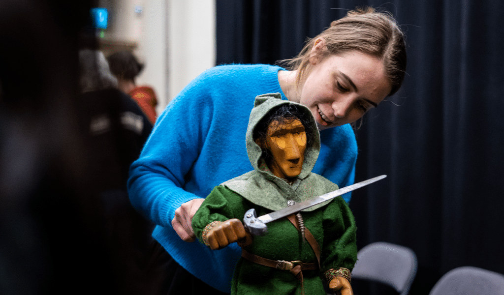 Exeter Northcott Futures Puppetry Masterclass promotional image. Background: Black theatre curtain drawn almost entirely across a white wall. A group of people out of focus. Foreground: A woman wearing a light blue jumper operates a rod puppet of Robin Hood, wearing a dark green tunic, light green hood and holding a sword.