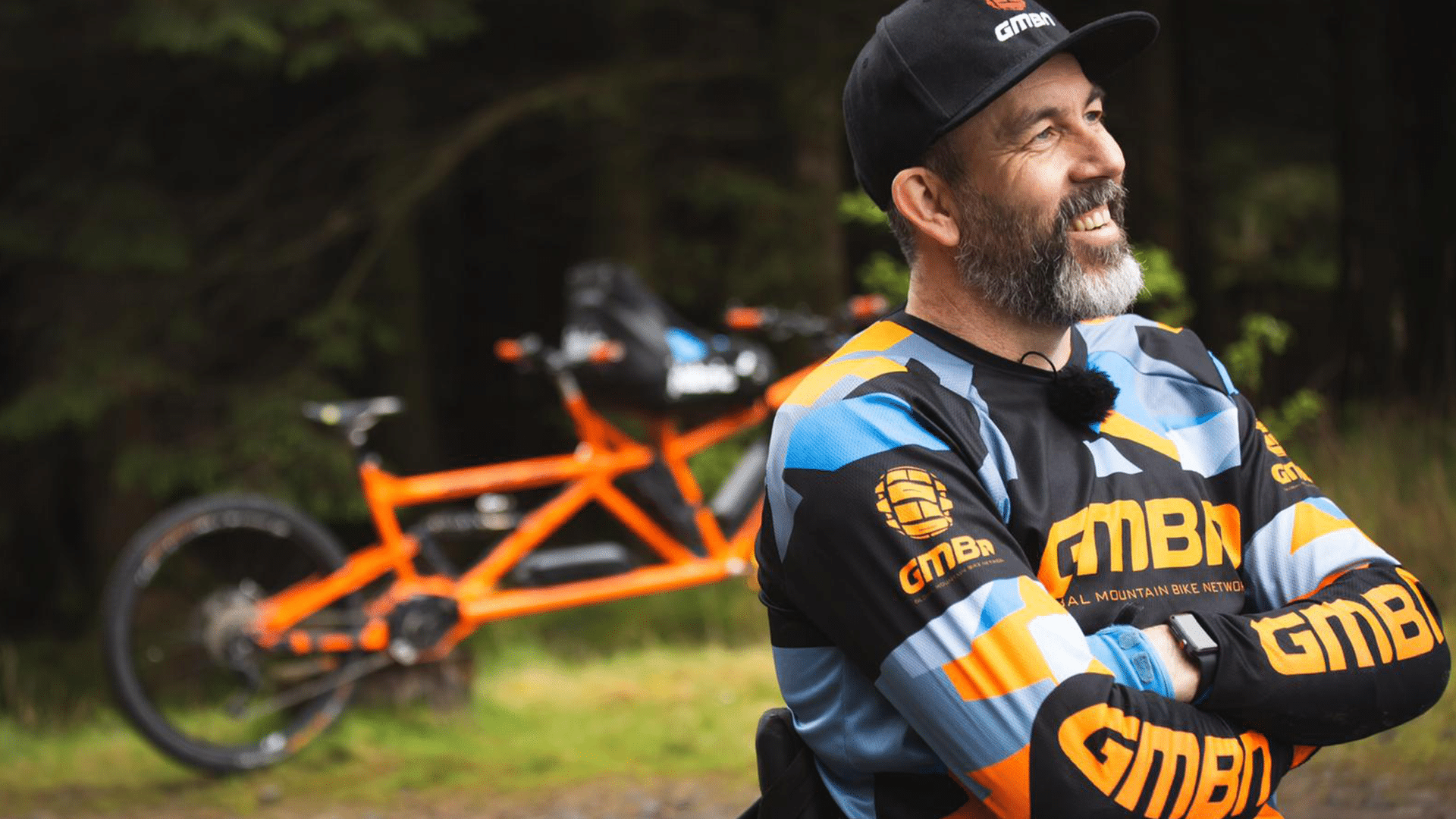 Martyn Ashton Bike Party promotional photo. Background: Forest. Tall trees. An orange coloured two person mountain bicycle, with the front of the bicycle adapted for a disabled rider. Foreground: (right) Martyn Ashton, wearing a black 'GMBN' hat and a black, blue and orange 'GMBN' mountain bike rider's jersey, crossing their arms and smiling.