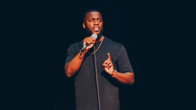 A photo of Mo Gilligan on stage wearing a black t-shirt and holding a microphone in his right hand and holding a finger up on his left hand. The background is black.