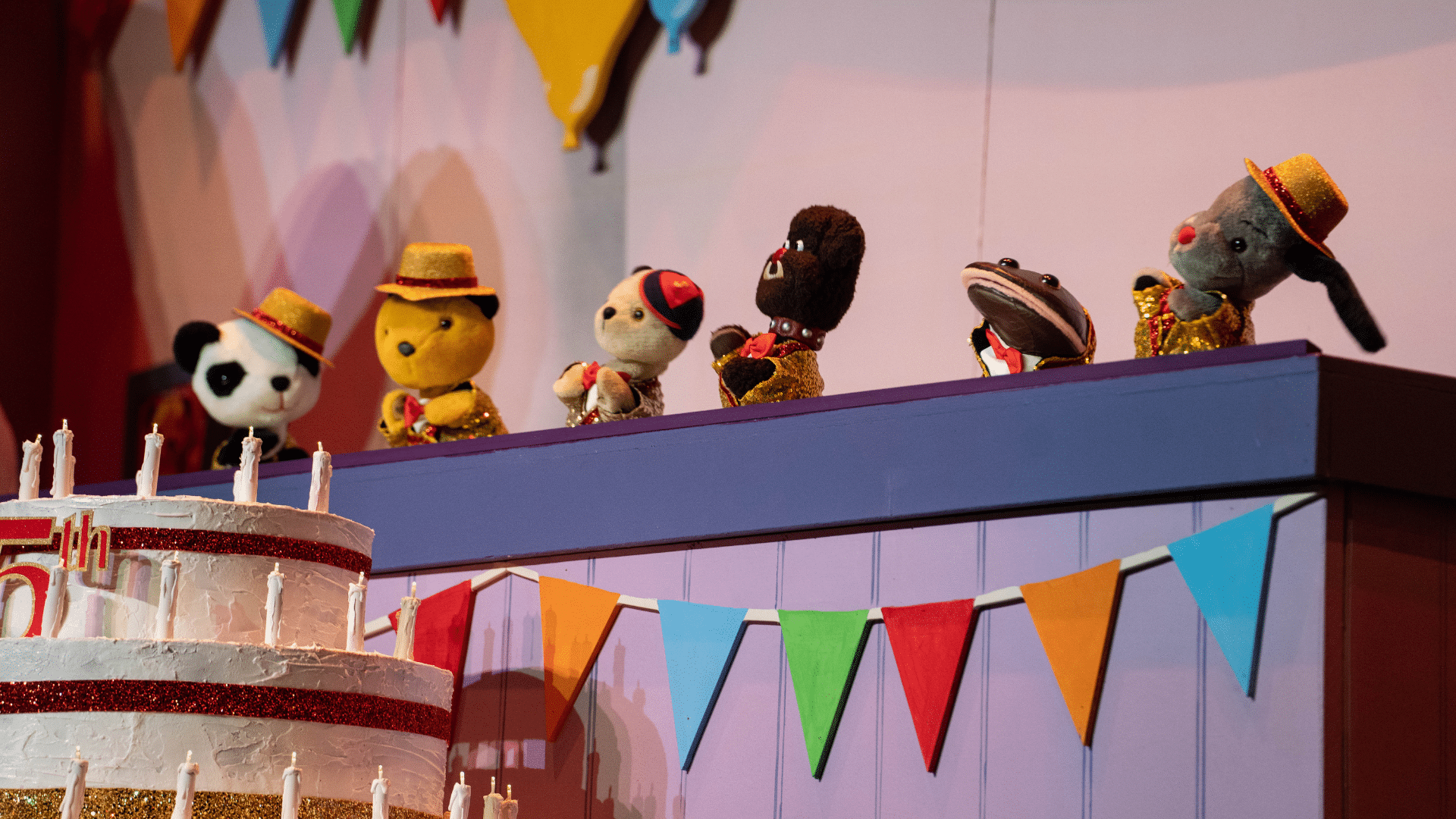 The Sooty Show production image: Sooty and his puppet friends look at a large cake with candles on it; there is bunting along the purple wooden walls.