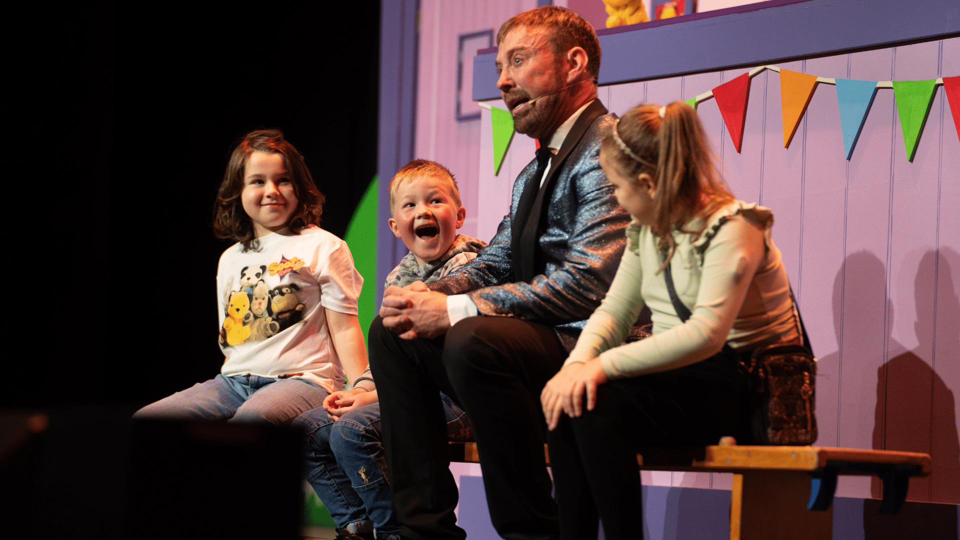 The Sooty Show production image: a man in a sparkly blue dinner jacket sits on a bench with three smiling children; behind them is a lilac wooden wall with colourful bunting.