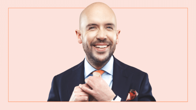 Image of Tom Allen adjusting his tie and smiling. He wears a smart suit. The background is a peach colour with an orange rectangular border.