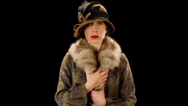 A Room of One's Own promotional photo - A photo of a woman wearing a fur-lined coat and a brown hat, holding the lining of her coat with her hands. The background is black.