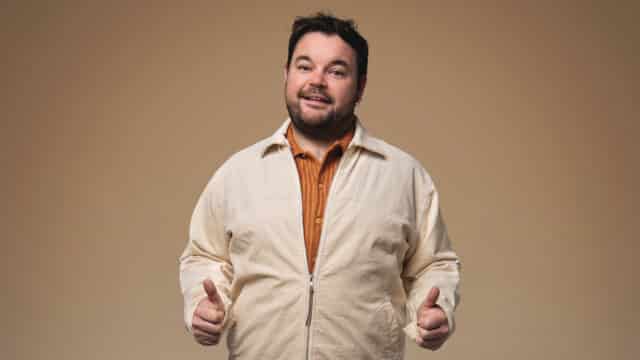 Promotional photo of Lloyd Griffith wearing a cream coloured jacket and an orange shirt, stood facing forwards with his thumbs up by his side, with a half-smile on his face.