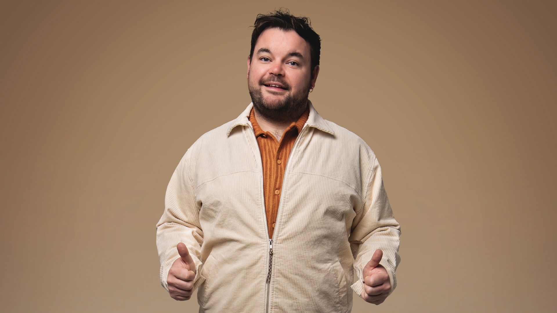 Promotional photo of Lloyd Griffith wearing a cream coloured jacket and an orange shirt, stood facing forwards with his thumbs up by his side, with a half-smile on his face.