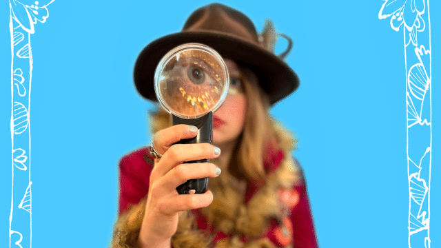A blonde woman with a wide-brimmed hat is looking at the camera through a magnifying glass