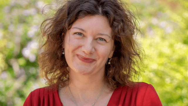 A headshot of Professor Turi King: a white woman with curly brown hair, wearing a red top and necklace; she is smiling; the background is green leaves.