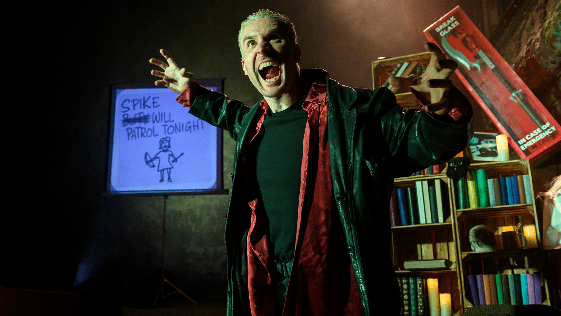 Buffy Revamped production photo - Brendan Murphy, his arms out wide holding his fingers like claws, his mouth wide open as if roaring. He wears an open red shirt underneath a black leather jacket. In the background there are bookshelves, a red 'break glass in case of emergency' case with an axe inside and a small screen with the words 'Spike will patrol tonight' handwritten with the word 'Buffy' crossed out where 'Spike' is. There is a crude childlike drawing of a person holding a bow and arrow underneath.