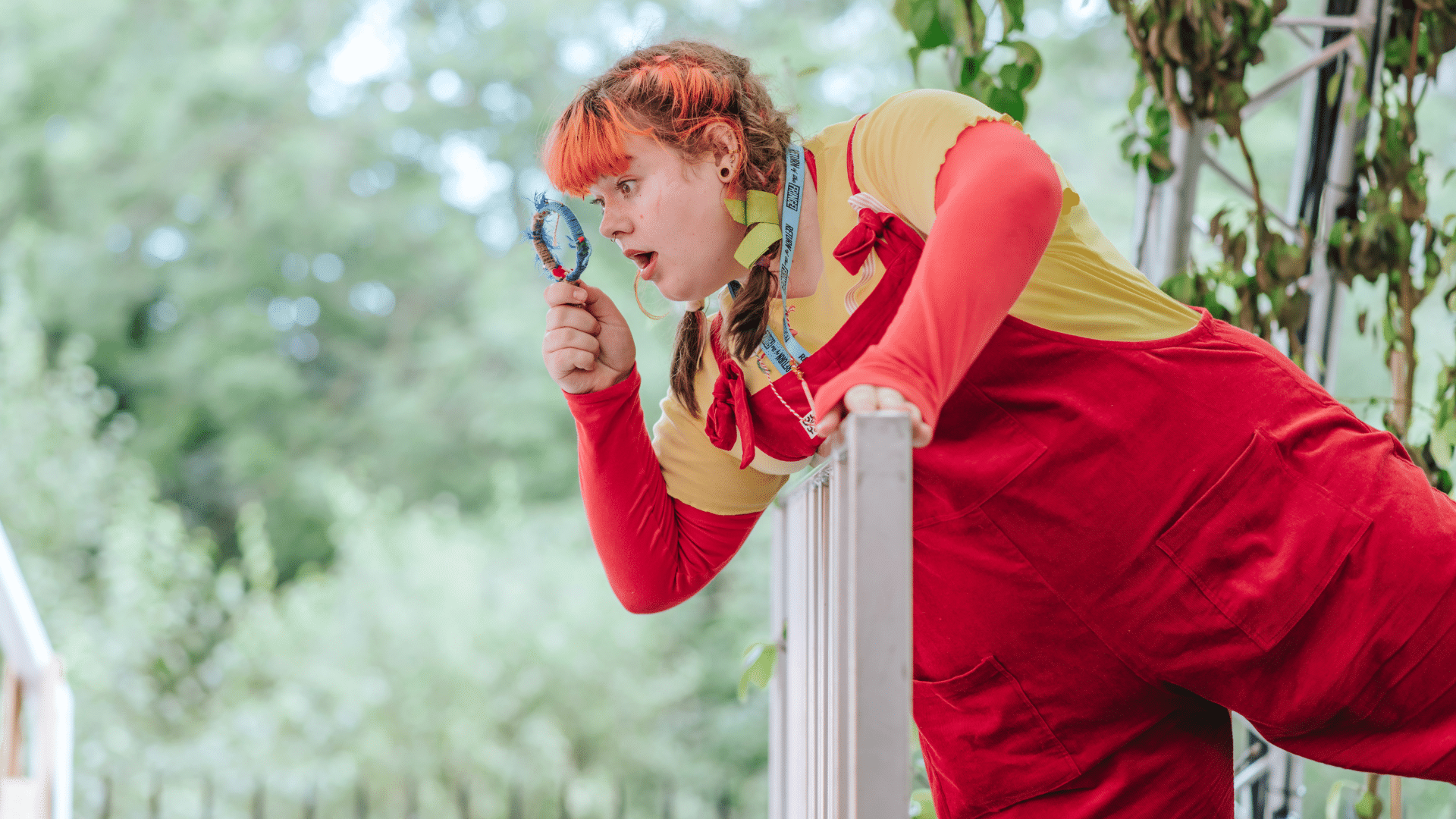 Meet Astrid Lindgren's Pippi Longstocking production photo - Sofie Miller as Pippi Longstocking holding a magnifying glass to her face and looking through it. She has red hair in pigtails and wears red dungarees above a yellow top.