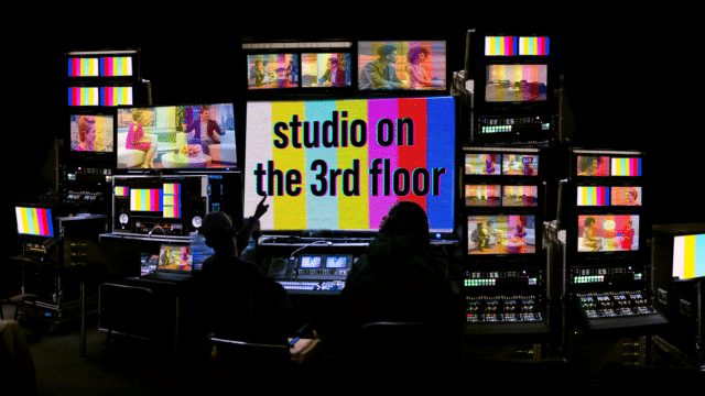 An image of a TV studio gallery, with two silhouetted people sat in front of several screens from small to large in size, and lots of technical equipment. The person on the left points at the largest screen in the middle which shows coloured bars and text reading 'studio on the 3rd floor'.