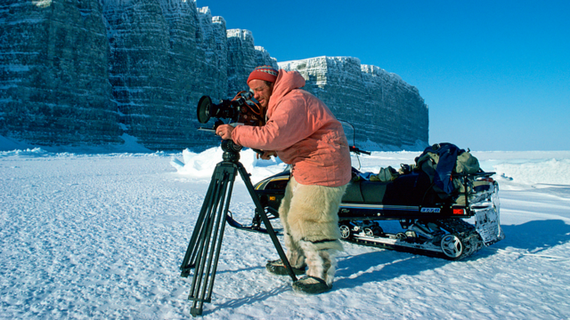Doug Allan in a snowy tundra environment, looking into the lens of a film camera on a tripod. Behind him is a snowmobile. Icy cliffs are in the background.