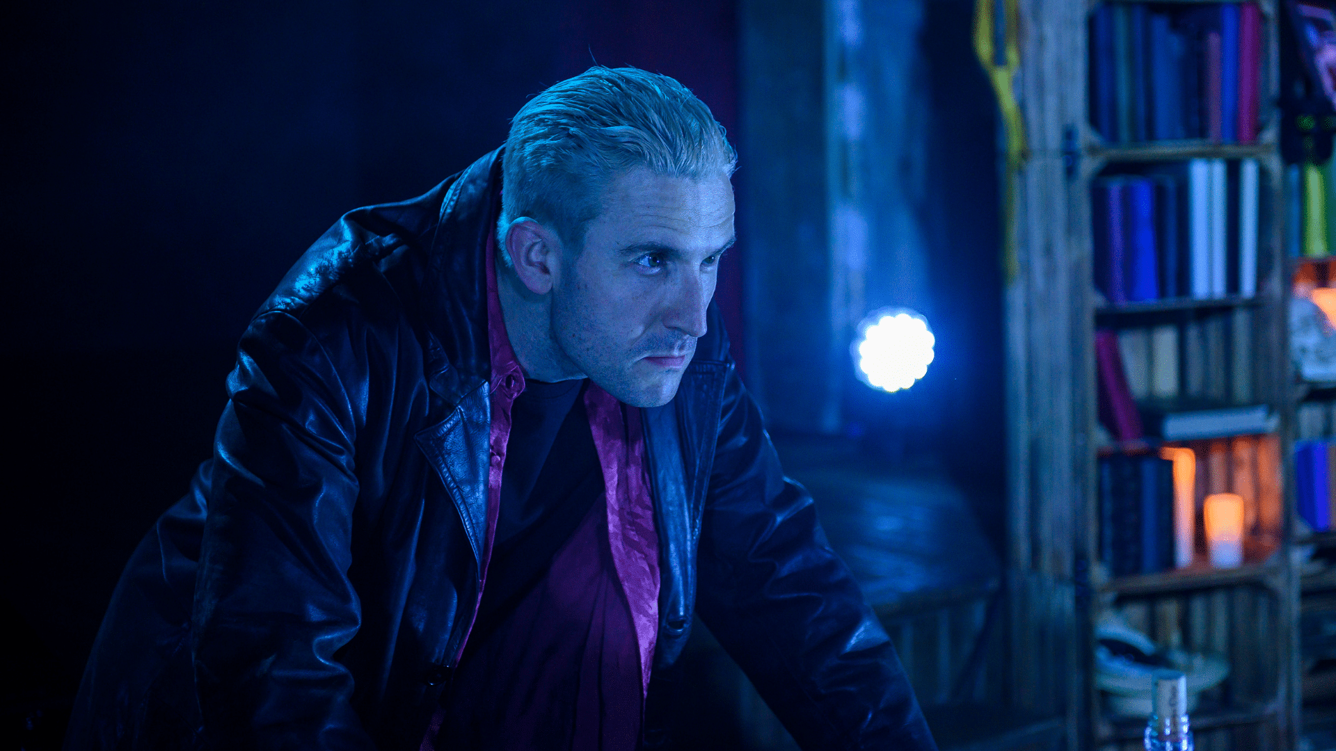 Buffy Revamped production photo - Brendan Murphy hunched over, looking to his left with a blank expression on his face. He is bathed in blue light. In the background there is a bookshelf.