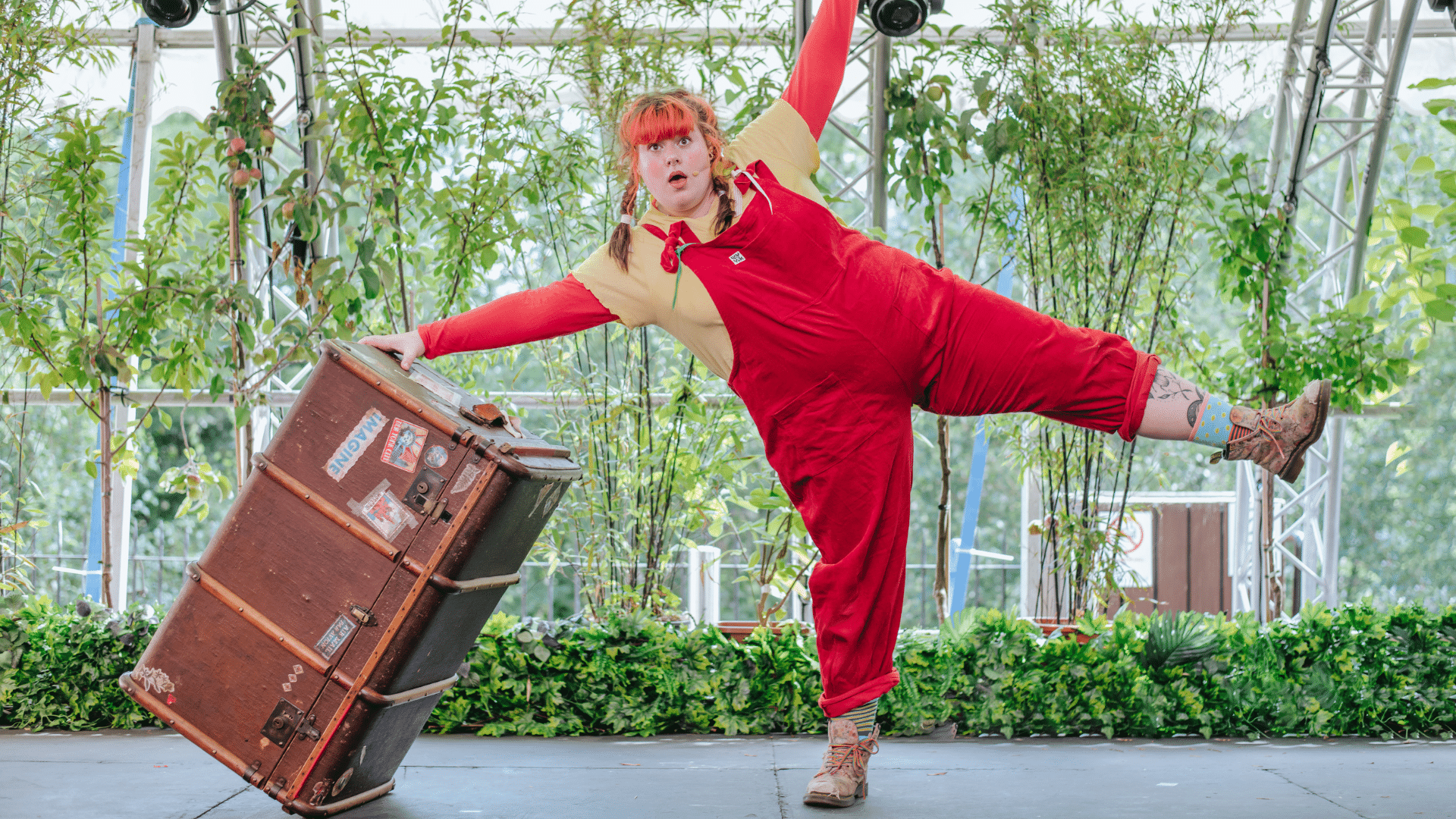 Meet Astrid Lindgren's Pippi Longstocking production photo - Sofie Miller as Pippi Longstocking in a star pose while leaning on a suitcase with one leg and arm in the air. She has red hair in pigtails and wears red dungarees above a yellow top.