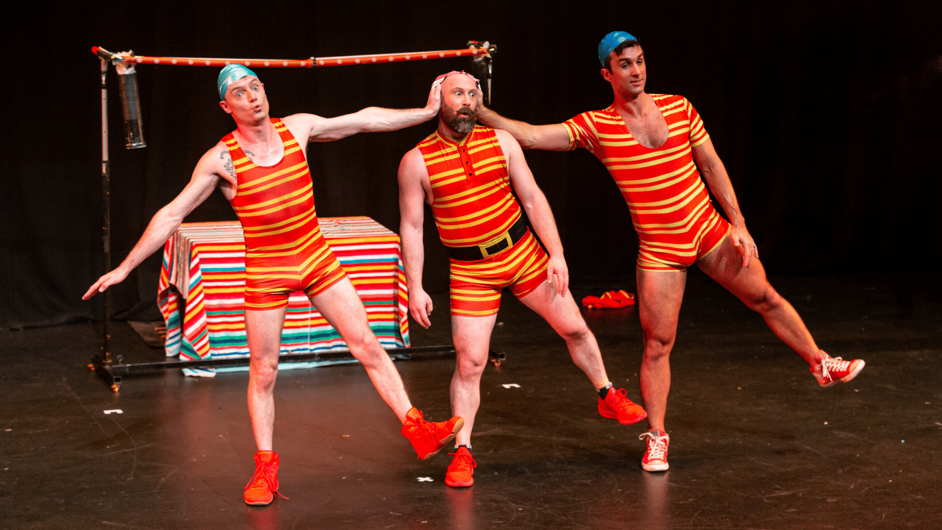 Splash Test Dummies production photo - The Dummies performing on stage, wearing old-fashioned striped bathing suits and swimming caps, stood in a line, each leaning to the side with one foot off the ground. The men on either side of the man in the middle have a hand each on the middle man's head.