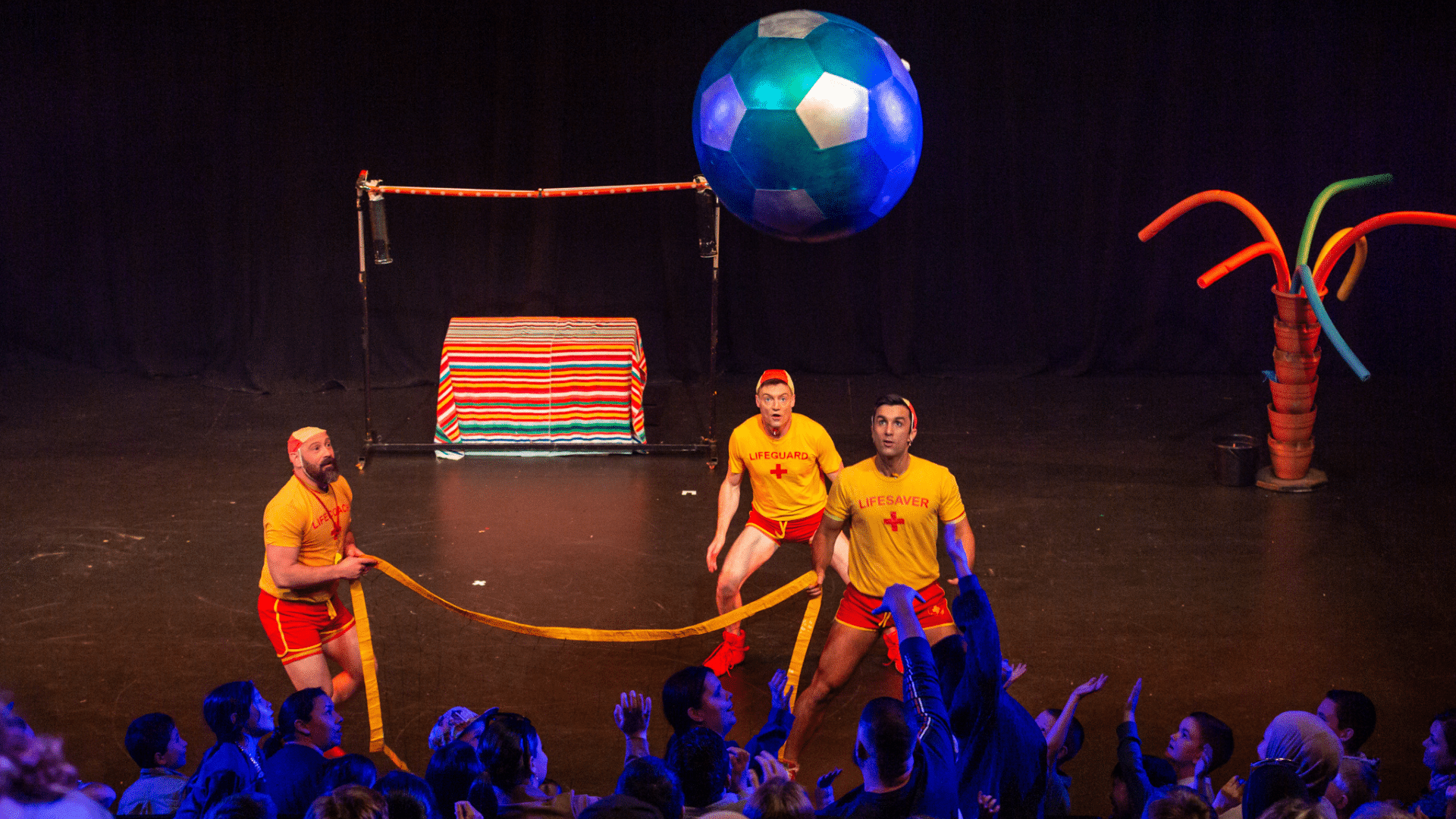 Splash Test Dummies production photo - The Dummies performing on stage, wearing yellow and red lifeguard outfits. They are stood in front of a packed crowd watching a giant beach ball in the air above the audience.