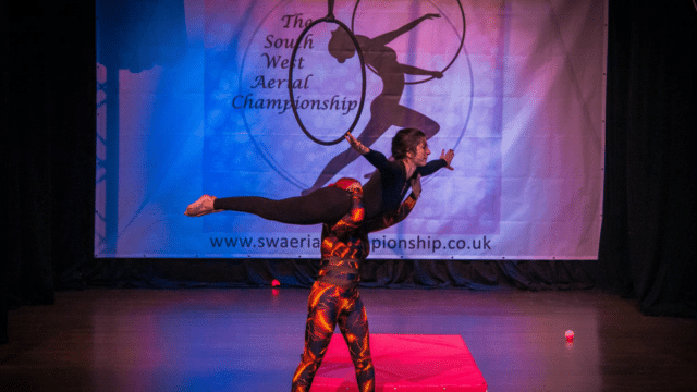 Aerial Allsorts: South West Aerial Championships promotional photo - A performer being lifted in the air by another performer. In the background: A banner with 'The South West Aerial Championship' logo.
