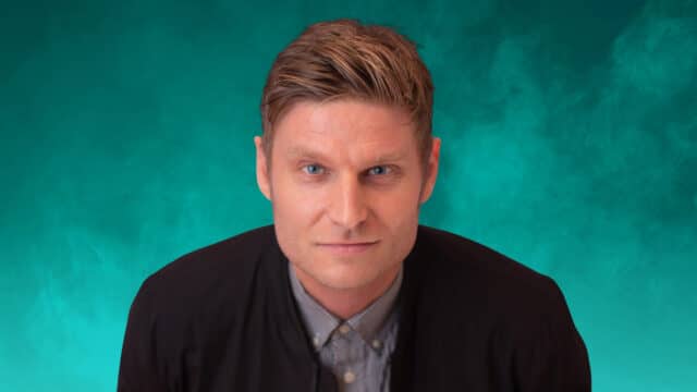 Promotional photo of comedian Scott Bennett - Scott Bennett looking forwards. The background is lit turquoise with swirly smoke.