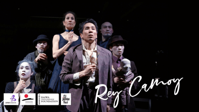A photo of members of the tarinainanika theatre company grouped together on stage with serious expressions on their faces. A man at the front holds a paint brush and a man to his left holds a bust with material over the face. Signature style text reads: Rey Camoy.