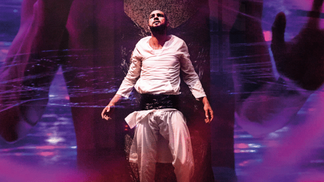 The Chosen Haram promotional artwork - a man in white robes with a large black belt around his waist, stood with his arms slightly out by his sides, staring upwards. Background: a giant figure looms, appearing to be behind a transparent barrier - chest, arms and a hand are visible. The image is bathed in a pink-purple light and swirly lines and shapes can be seen blended into the image.