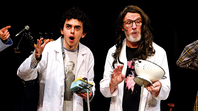 The Washing Machine of Destiny production photo. Black background. Two men wearing white lab coats stand beside each other. The man on the left is young and is holding a musical pad instrument with a white ringed telephone cord coming out of it. The man on the right is older, wearing a brown wig, and is holding a white colander with a white ringed telephone cord coming out of it.