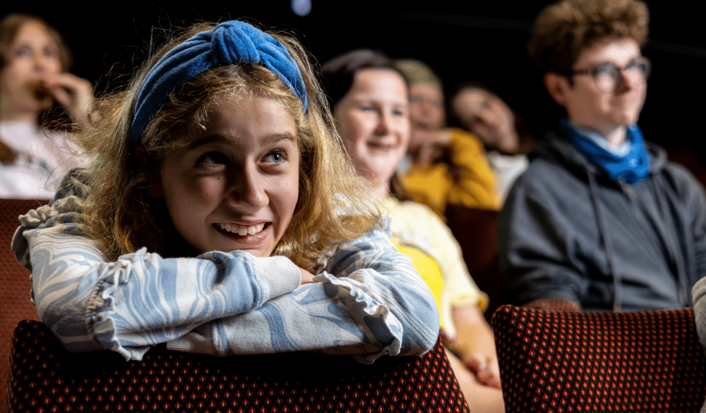 A young girl wearing a blue and white-coloured hoodie leans forward onto a red theatre seat. She is resting he head on her crossed arms, and is looking up and smiling at what is happening off-camera on stage. She is surrounded by other young people, also engaged with what is happening off-camera on stage.