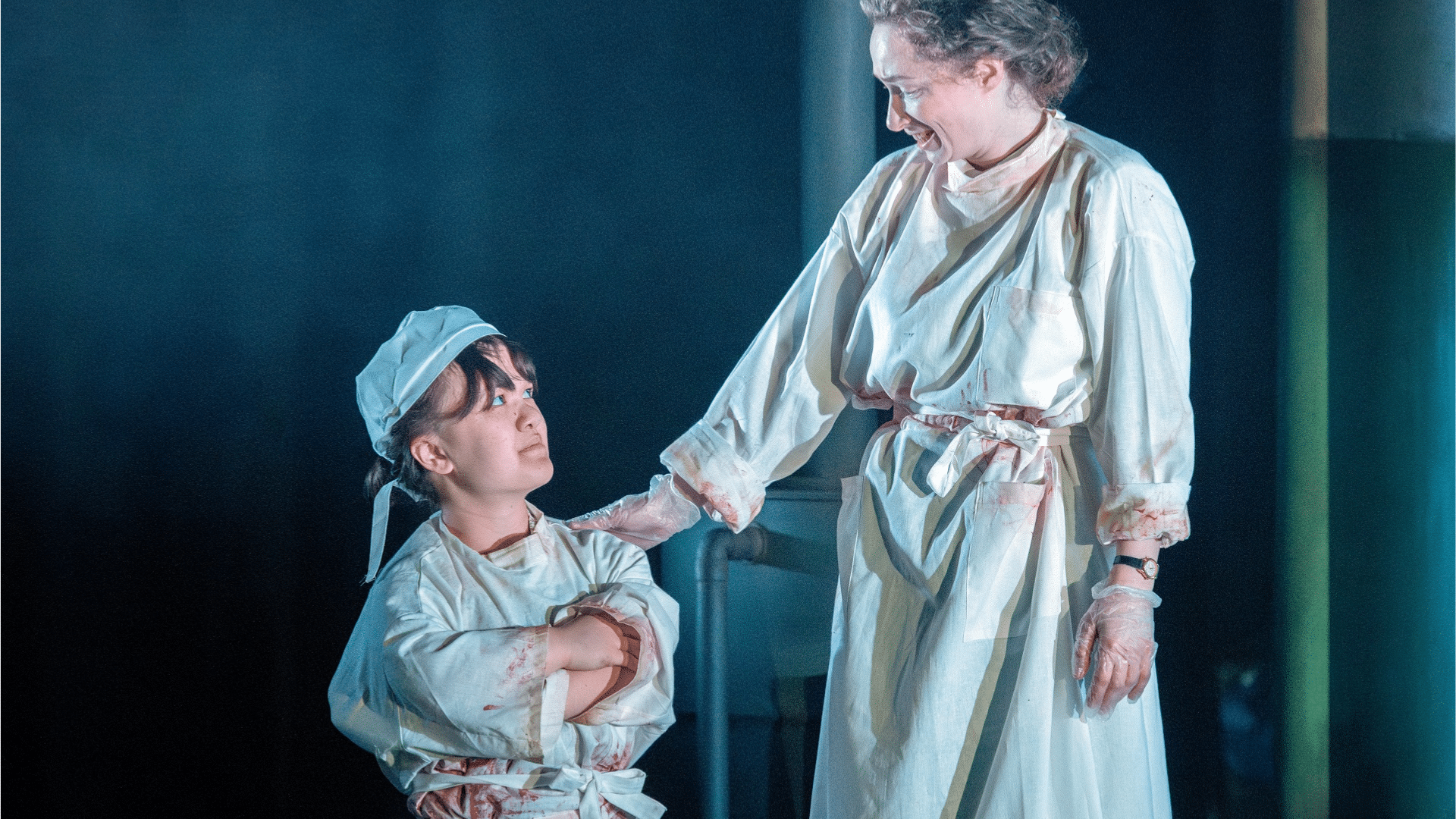 Frankenstein production photo: Annette Hannah and Eleanor McLaughlin (playing Victoria Frankenstein) are both dressed in white surgical scrubs, with small blood splatters. Hannah (left) is crossing her arms and looking up at McLaughlin, who smiles and grasps her shoulder.