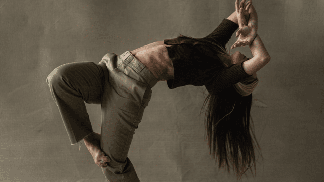 Richard Chappell Dance Home: Revisited promotional photo. A woman wearing a brown top and beige trousers leans back in the air hair falling down with her arms crossed as she balances standing on her left leg as her right leg wraps rests on her left knee.