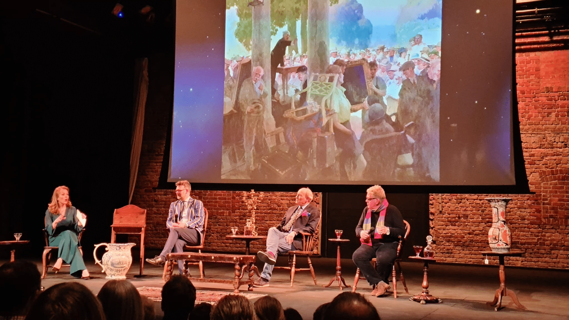 Foreground: silhouette of audiences' heads. Midground: Charlie Ross, Christina Trevanion, Charles Hanson and Phil Serrell sitting on chairs on a stage surrounded by a selection of antique items. Background, a large projection screen showing a photograph of a painting.