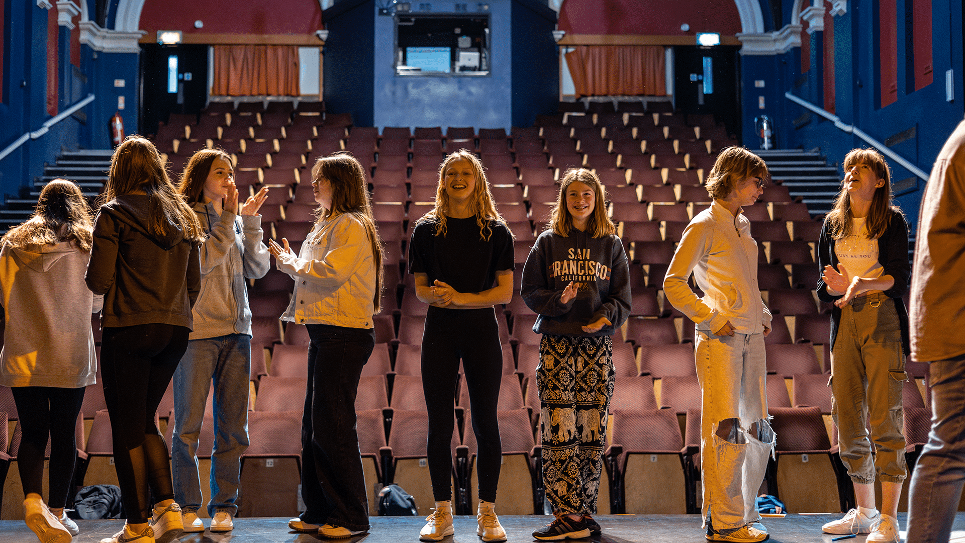 Barnfield Theatre auditorium. A mixed group of young people talk to each other while standing on the Barnfield Theatre auditorium stage. They have their backs turned to the auditorium seats, which are empty.